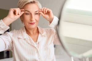 Mature woman performing face-building exercises near a mirror in her bedroom, focusing on a natural brow lift and facial toning.